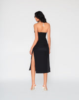 Rear view of a woman modeling the black Cate midi dress, highlighting its open back design, white halter tie cords, and high thigh high side slit.