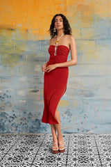 Full length lifestyle portrait of a woman standing against a multi-colored brick wall, wearing the red Cate Ruched Halter Dress with white strappy platform heels.