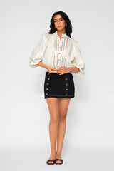 Full-length studio image of a woman modeling the ivory Eva cotton lace blouse paired with a high-waisted black mini skirt and brown slides.