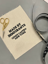 Overhead shot of a canvas fabric swatch embroidered with "MADE BY IMMIGRANTS CINDY CASTRO NEW YORK", positioned next to golden tailoring shears and a measuring tape.
