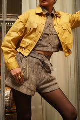 Photo of a woman from the chin down, modeling the cropped marigold yellow Frida cotton twill jacket, styled with high-waisted tweed utility shorts and black tights.