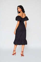 Rear studio shot of a woman modeling the black Gabriela cotton dress, showcasing its elegant off-the-shoulder back neckline and structured midi skirt with a gathered hem ruffle.