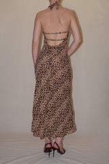 Rear studio perspective of a model showcasing the dramatic low open-back design, delicate horizontal string straps, and halter neck tie on the leopard print Jolene maxi dress.