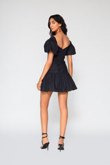 Rear studio shot of a woman modeling the black Manuelita mini dress, highlighting its elegant low V-back design and flouncy gathered skirt line.
