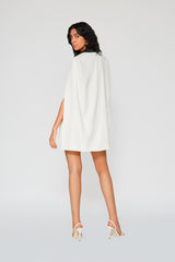 Full-length rear view of a model wearing the ivory Michelle Blazer Dress, showcasing the fluid draping of the cape design and sleek, minimal back silhouette paired with white stiletto heels.