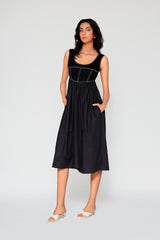 Full length studio portrait of a woman wearing the black Monica Denim Midi Dress, showcasing its contrast topstitching, corset-style structured bodice, functional side pockets, and white kitten heel slides.