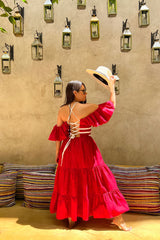 Rear lifestyle view of a model holding a sun hat, highlighting the intricate white lace-up corset back and off-the-shoulder styling option of the red cotton Norma dress.