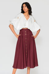 Full-length studio portrait of a woman modeling the dark burgundy Patricia linen midi skirt with a button-down front placket and a gathered A-line skirt.