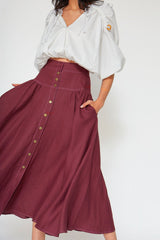Dynamic close-up shot of a model with her hand in her pocket, showcasing the fluid drape, side pockets, and white contrast topstitching of the burgundy linen midi skirt.
