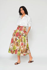 Full-length studio image of a woman modeling the high-waisted Patricia printed long skirt, featuring a vibrant watercolor floral pattern in red, green, and tan, styled with a white button-down shirt.