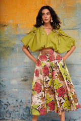 Image of a model standing against a distressed blue wall, wearing the olive green Zoila ruched top paired with a colorful watercolor floral midi skirt.