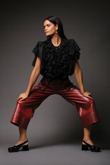 Full-length editorial portrait of a woman in a wide dynamic stance, modeling the black Miranda Chiffon Blouse featuring a cascading ruffled front panel and burgundy leather culottes.