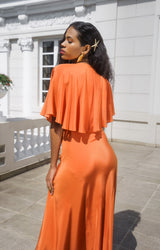 Rear outdoor perspective of a model highlighting the fluid satin drape of the skirt and the clean, circular look of the ruffle cape on the orange Natalia dress.