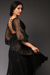 Close-up side-rear view of a model presenting the sheer texture of the voluminous sleeves and the low-cut, criss-cross strap detailing on the back of the black and gold Paula dress.