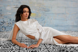 Model reclining in the Alicia white silk midi dress featuring draped cape sleeve, side cutout, front slit, and luxurious satin fabric.