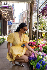Model wearing the Cameron yellow bubble mini dress beside colorful flowers featuring puff sleeves, side cutouts, and voluminous bubble skirt.