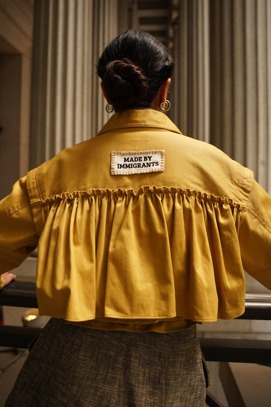 Rear view of a woman wearing the marigold yellow Frida Twill Jacket, highlighting its dramatic gathered ruffle back yoke and a stitched patch reading "MADE BY IMMIGRANTS".
