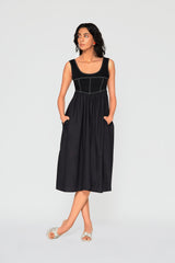 Full-length studio profile of a model standing with hands in her pockets, displaying the natural drape of the gathered A-line skirt on the black Monica denim dress.