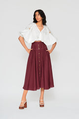 Full-length studio image of a model wearing the high-waisted burgundy Patricia linen midi skirt, paired with a voluminous white cropped top and platform heels.
