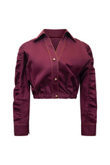 Studio product view displaying the front profile of a plum violet cropped jacket shirt, showcasing its structured collar, button-down front with gold metallic hardware, contrasting topstitching, and heavily ruched sleeves.