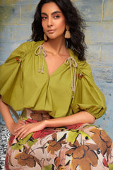 Close-up shot of a model sitting, highlighting the olive green Zoila blouse's ruched shoulder details, keyhole ties with wooden accents, and flowing cotton fabric.