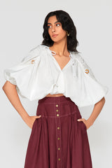 Studio portrait of a woman wearing the white Zoila Cotton Ruched Top, showcasing its V-neckline, white cord details with wooden rings, and voluminous puff sleeves styled with a burgundy skirt.