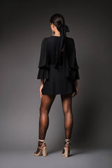 Rear studio perspective of a model highlighting the loose, fluid drape, long neck scarf tie, and tiered ruffle sleeves on the back of the black Jean silk dress.
