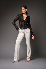 Full-length studio portrait of a woman modeling the black Joan Sheer Lace Top with a plunging satin-trimmed V-neckline, styled with tailored high-waisted white trousers.
