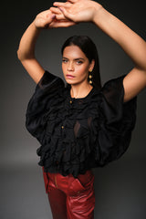 Three-quarter studio portrait of a model with arms raised, highlighting the dense tiers of sheer vertical ruffles, gold button hardware, and voluminous flutter sleeves of the black Miranda blouse.